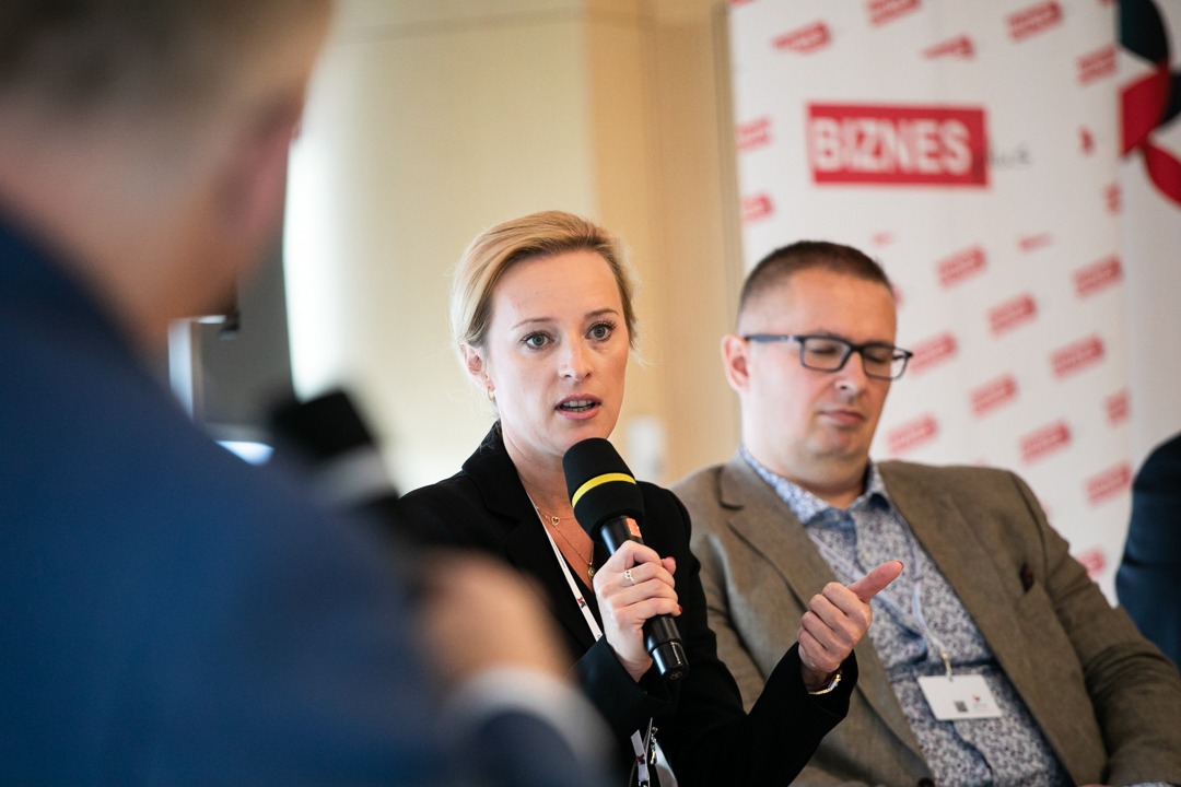 Kobieta w czarnej marynarce przemawia do mikrofonu podczas konferencji biznesowej, w tle mężczyzna w brązowej marynarce i logo firmy.