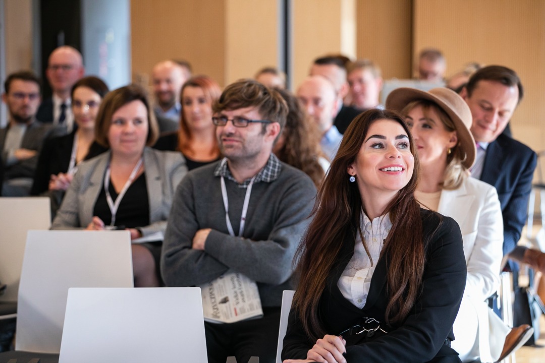 Uczestnicy konferencji siedzą rzędami na krzesłach, skupieni na prelegencie, z uśmiechniętą kobietą w czarnym żakiecie na pierwszym planie.