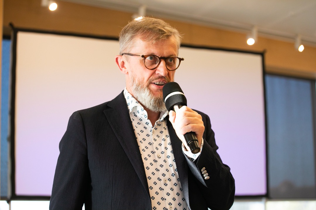 Mężczyzna w okularach i marynarce przemawia do mikrofonu na tle ekranu projekcyjnego.