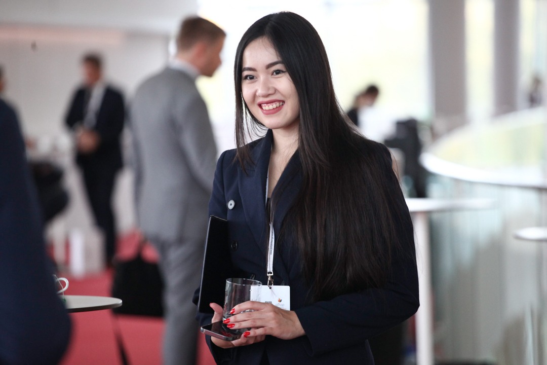 Uśmiechnięta kobieta w granatowym garniturze trzymająca szklankę i telefon na evencie, widoczne osoby w tle.