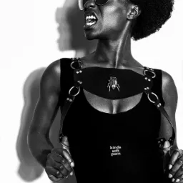 Czarno-białe zdjęcie kobiety w okularach przeciwsłonecznych z otwartymi ustami i wyrazistym cieniem na ścianie, ubrana w czarny strój z ozdobnym paskiem na piersiach i broszką w kształcie chrząszcza.