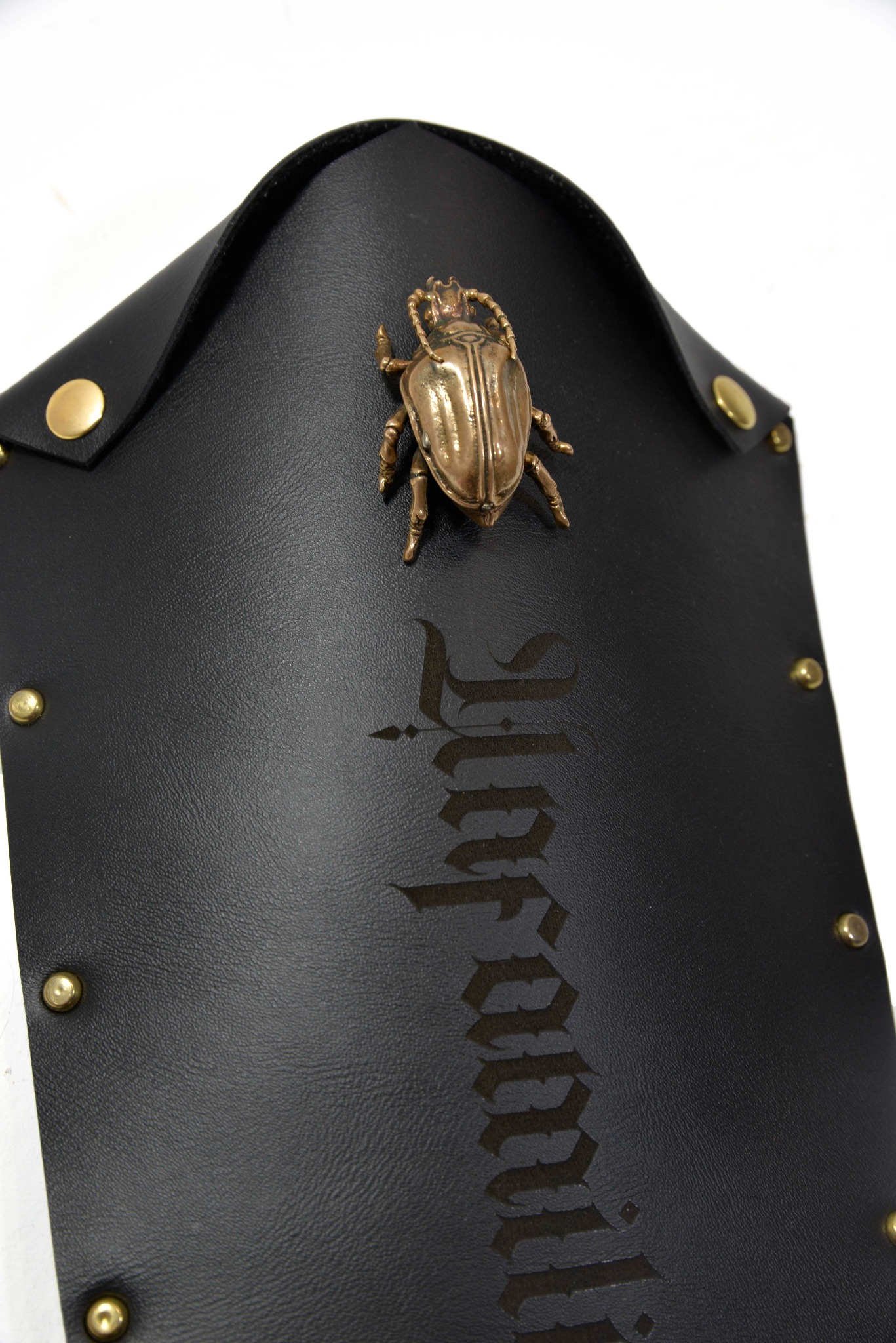 Czarna skórzana osłona z metalowym skarabeuszem i wygrawerowanym napisem w gotyckim stylu, zdobiona złotymi nitami.