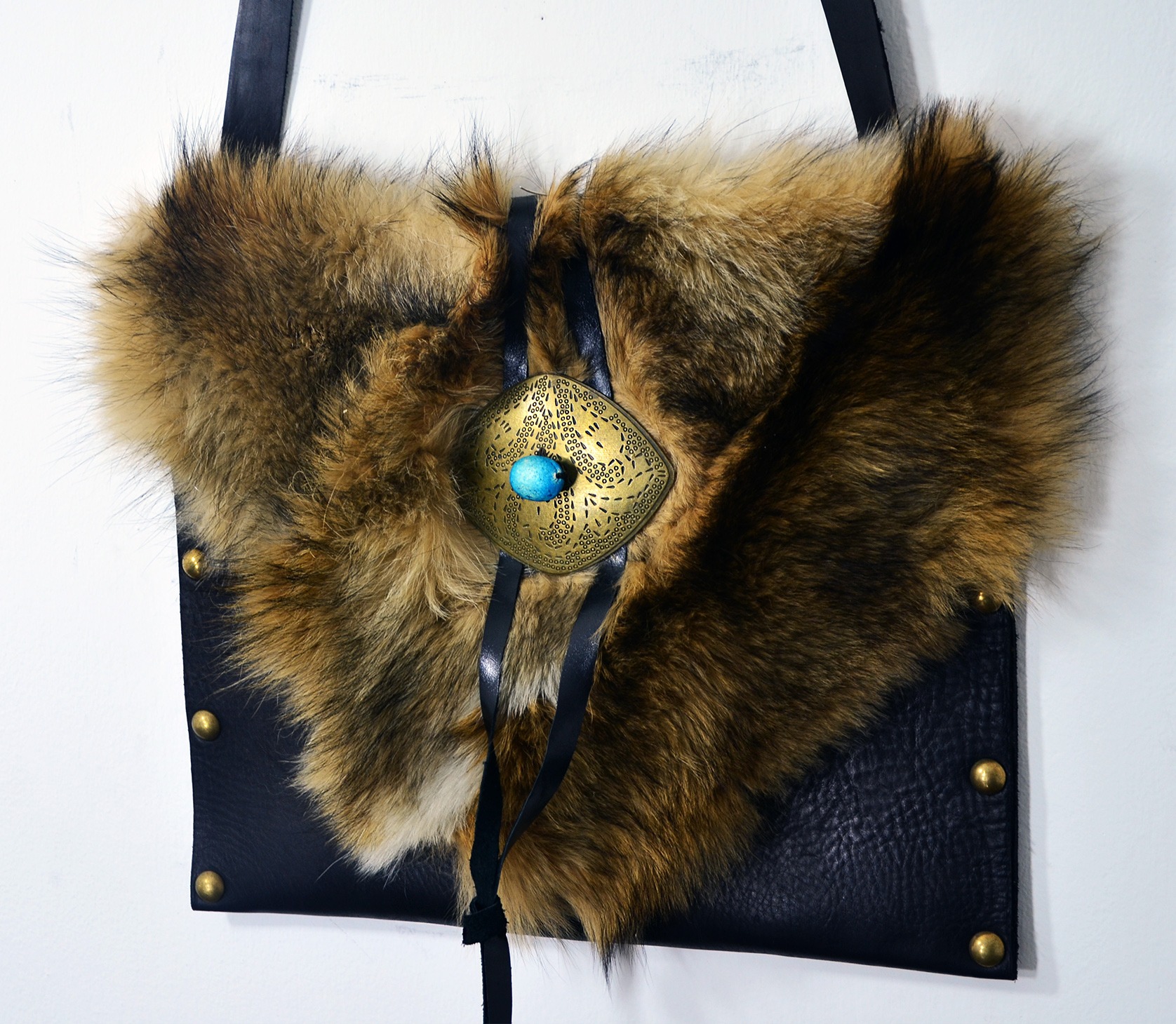 Ręcznie robiona torebka z czarnej skóry z futrzanym akcentem i ozdobnym, ażurowym, metalowym zapięciem z turkusowym kamieniem, zawieszona na skórzanym pasku.