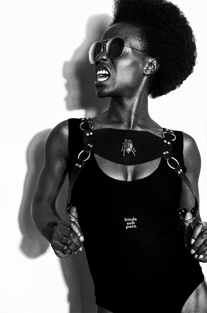 Czarno-białe zdjęcie kobiety w okularach przeciwsłonecznych z otwartymi ustami i wyrazistym cieniem na ścianie, ubrana w czarny strój z ozdobnym paskiem na piersiach i broszką w kształcie chrząszcza.