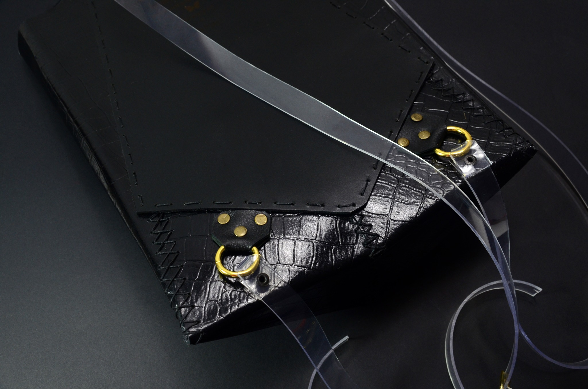 Czarna, skórzana torebka z fakturą skóry krokodyla, z przezroczystymi paskami i złotymi elementami, leżąca na czarnym tle.