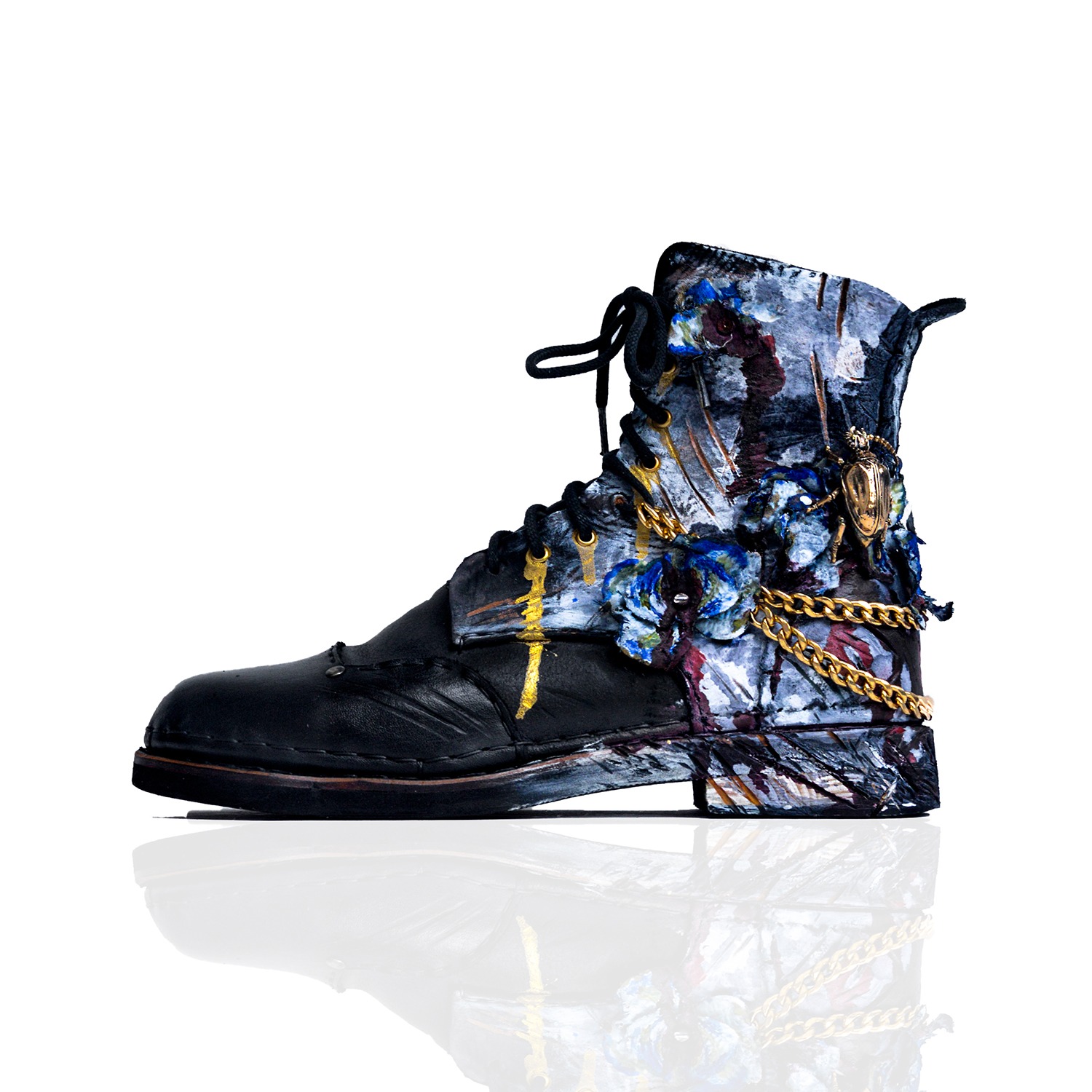 Czarny skórzany but typu worker, ręcznie malowany w abstrakcyjne wzory w odcieniach szarości, błękitu i złota, ozdobiony złotym łańcuchem i dekoracją w kształcie żuka, na białym tle z odbiciem.