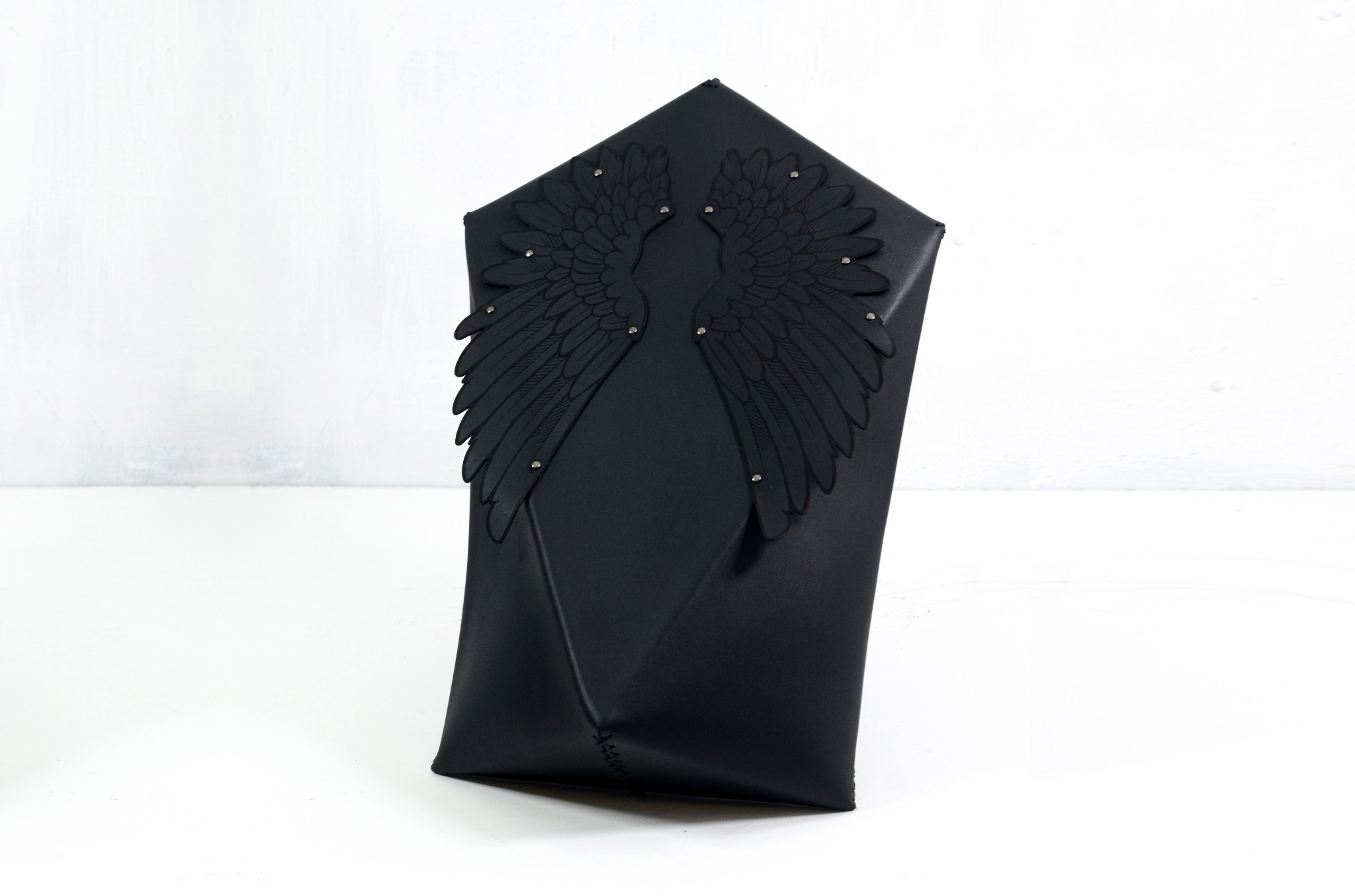 Czarna, skórzana torebka o geometrycznym kształcie z przyszytymi skrzydłami anioła ozdobionymi srebrnymi nitami, na białym tle.