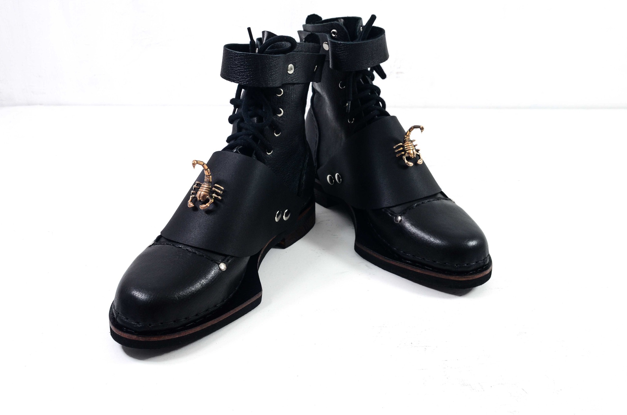 Para czarnych skórzanych butów typu workery z brązowymi ozdobami w kształcie skorpionów, widoczne sznurowadła i paski z metalowymi oczkami, na białym tle.