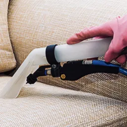 Dysza ekstraktora w różowej rękawicy czyści beżową tapicerkę sofy, widoczny niebieski wąż i czarne elementy urządzenia.