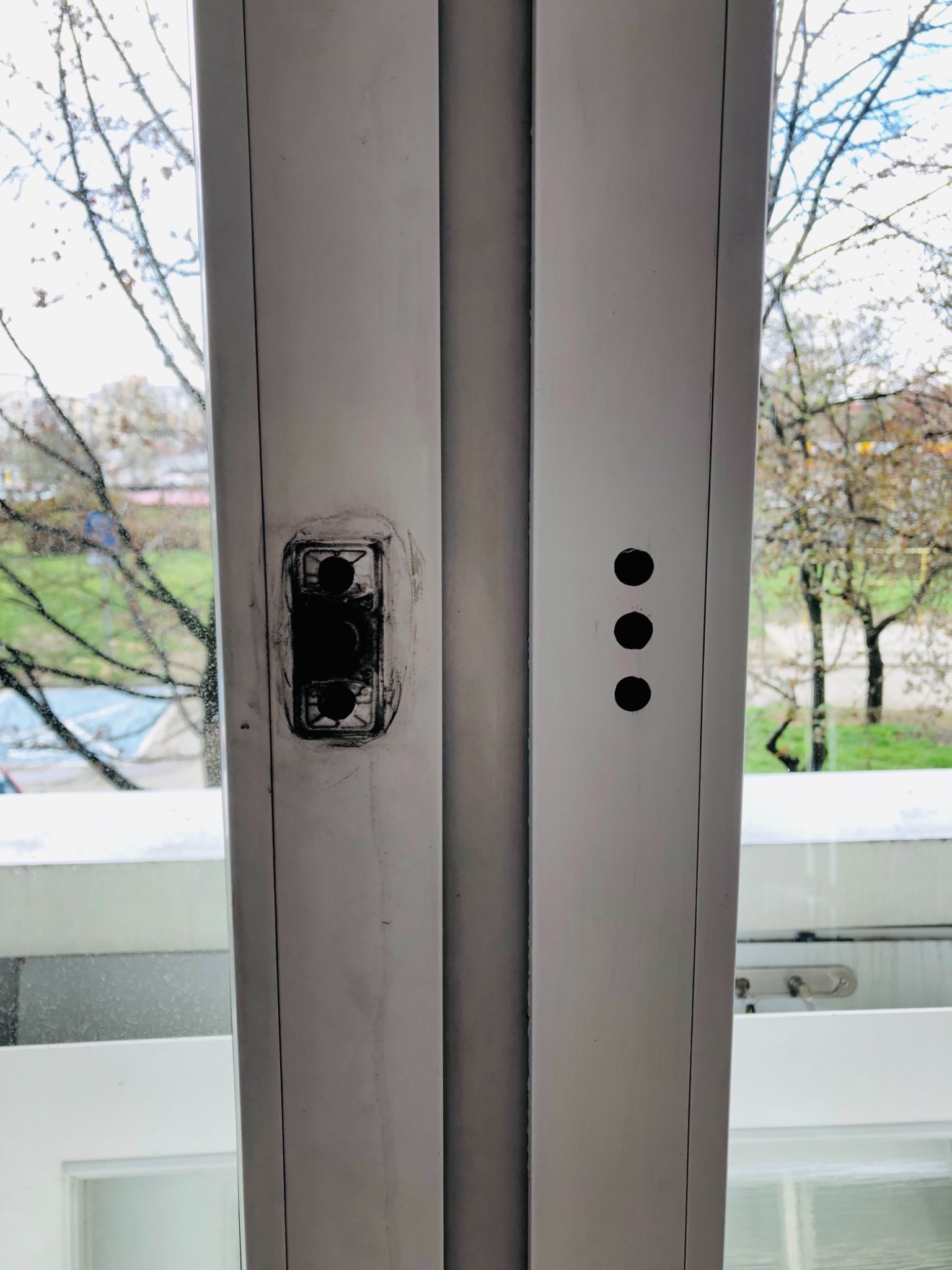 Biała rama okienna z widocznymi śladami po demontażu zamka i po wierceniu otworów, widok na zewnątrz przez szybę.