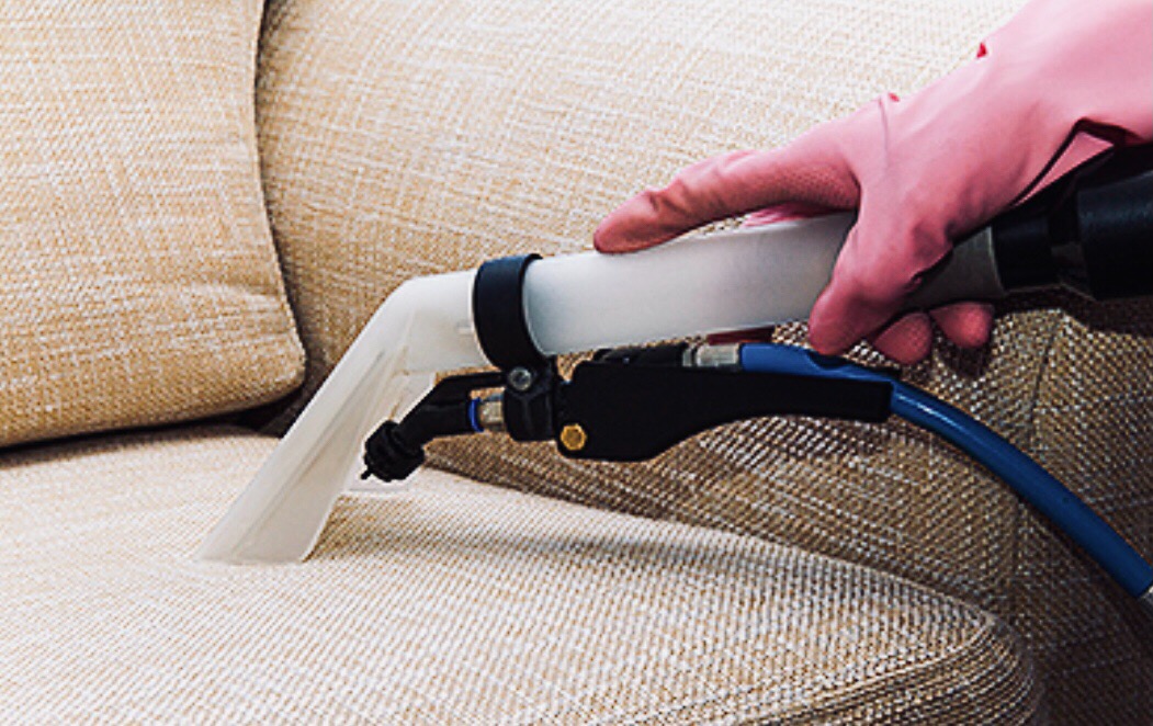 Dysza ekstraktora w różowej rękawicy czyści beżową tapicerkę sofy, widoczny niebieski wąż i czarne elementy urządzenia.