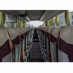 Wnętrze autokaru Mercedes Tourismo z beżowo-bordowymi fotelami z logo Mercedesa, widok z perspektywy pasażera w kierunku przodu pojazdu, z widocznymi zasłonkami w oknach.