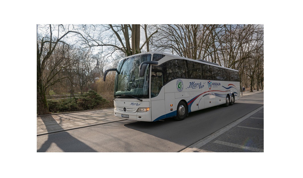 Biały autokar Mercedes Tourismo z logo firmy 'Marcin' Koszalin na tle parku miejskiego w słoneczny dzień.
