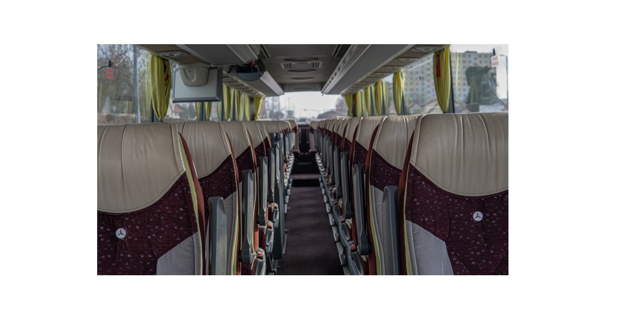 Wnętrze autokaru Mercedes Tourismo z beżowo-bordowymi fotelami z logo Mercedesa, widok z perspektywy pasażera w kierunku przodu pojazdu, z widocznymi zasłonkami w oknach.