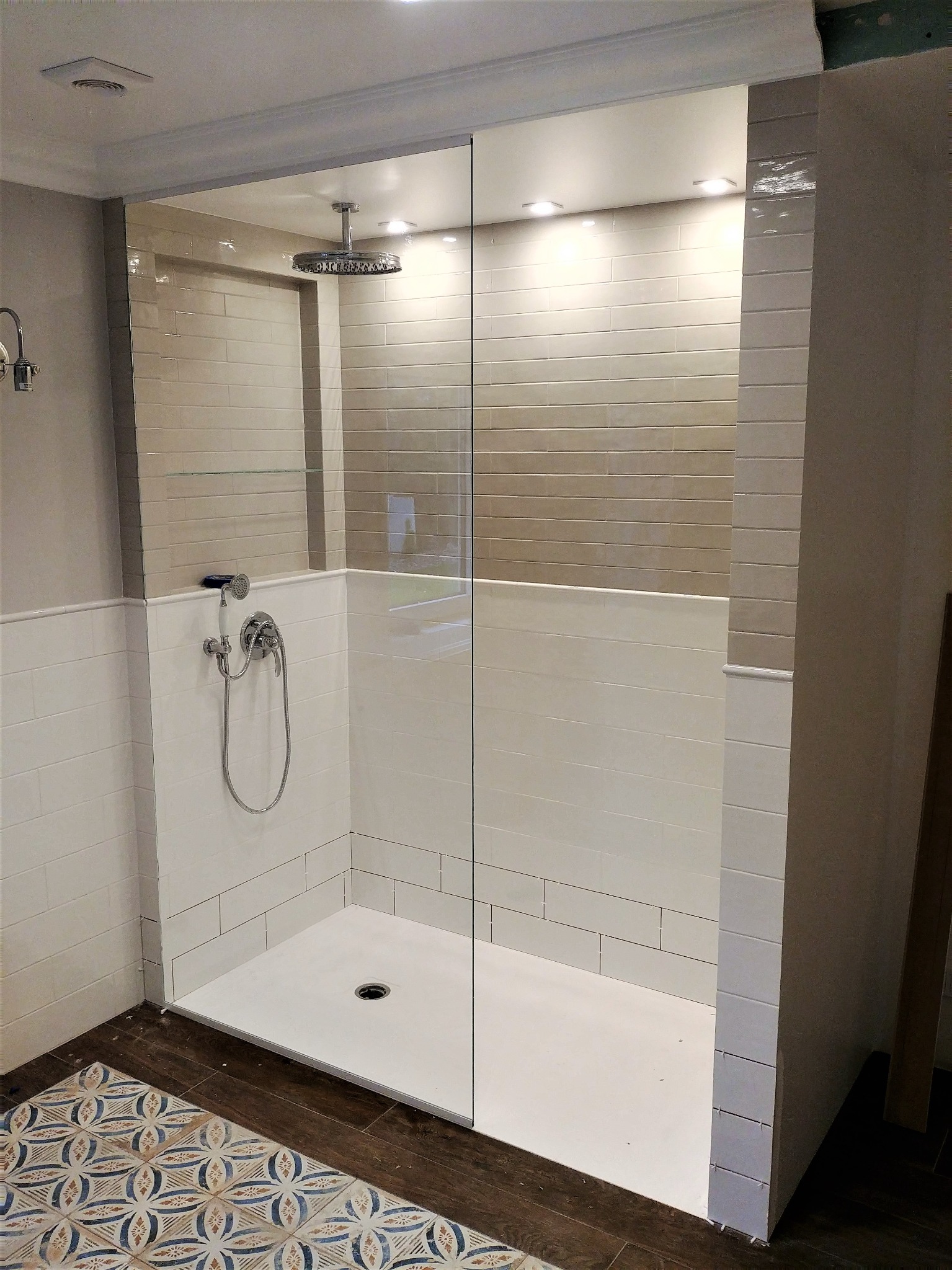 Nowoczesna kabina prysznicowa walk-in z chromowaną deszczownicą i baterią, wykończona białymi i beżowymi płytkami imitującymi cegłę, z dekoracyjną podłogą.