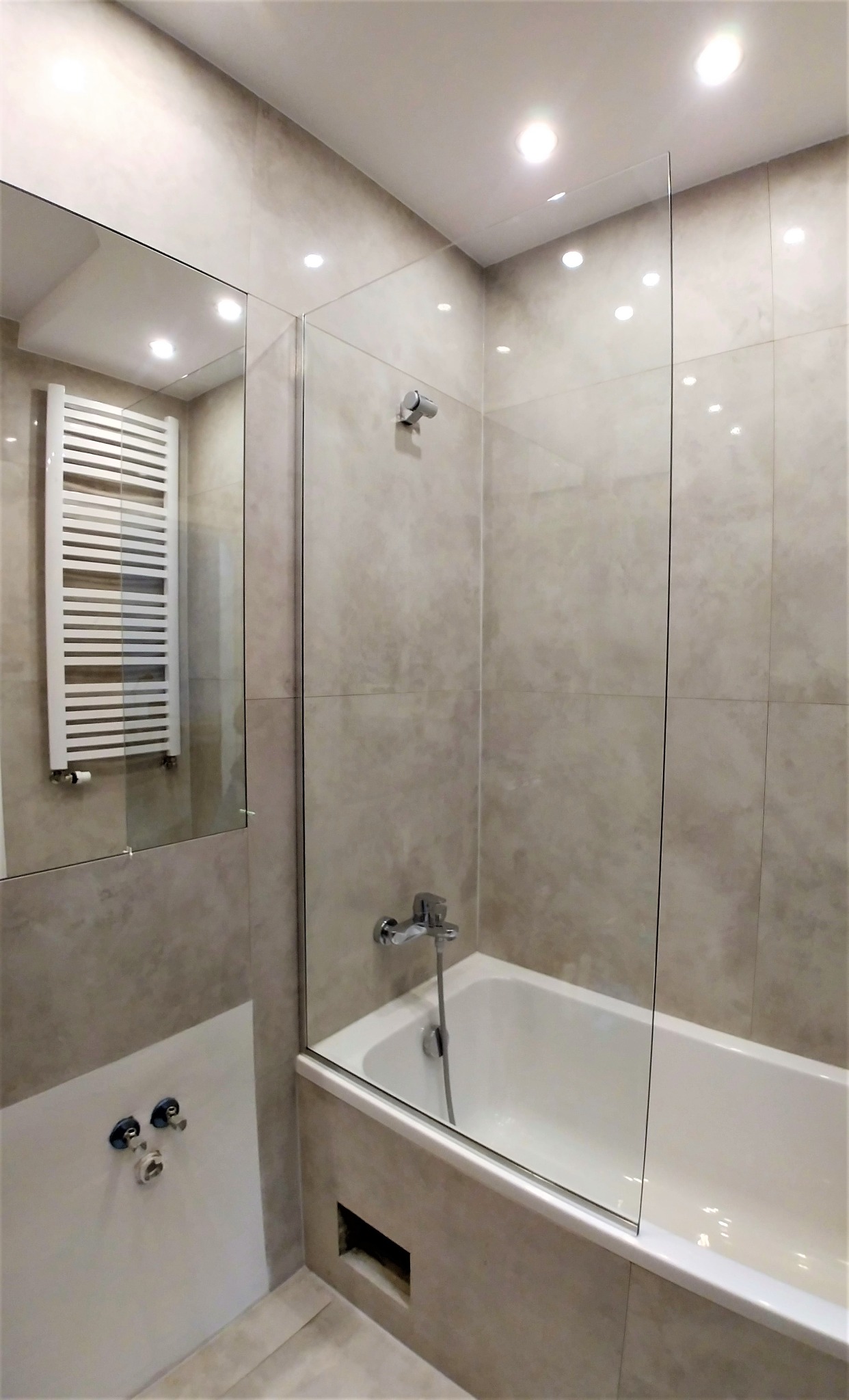 Nowoczesna łazienka z wanną, szklanym panelem prysznicowym i grzejnikiem drabinkowym odbijającym się w lustrze.