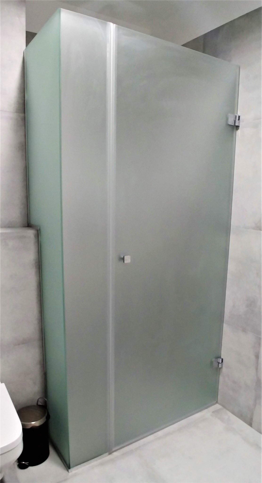 Kabina prysznicowa z matowego szkła, z widocznymi chromowanymi zawiasami i minimalistycznym uchwytem, w łazience wykończonej szarą płytką.