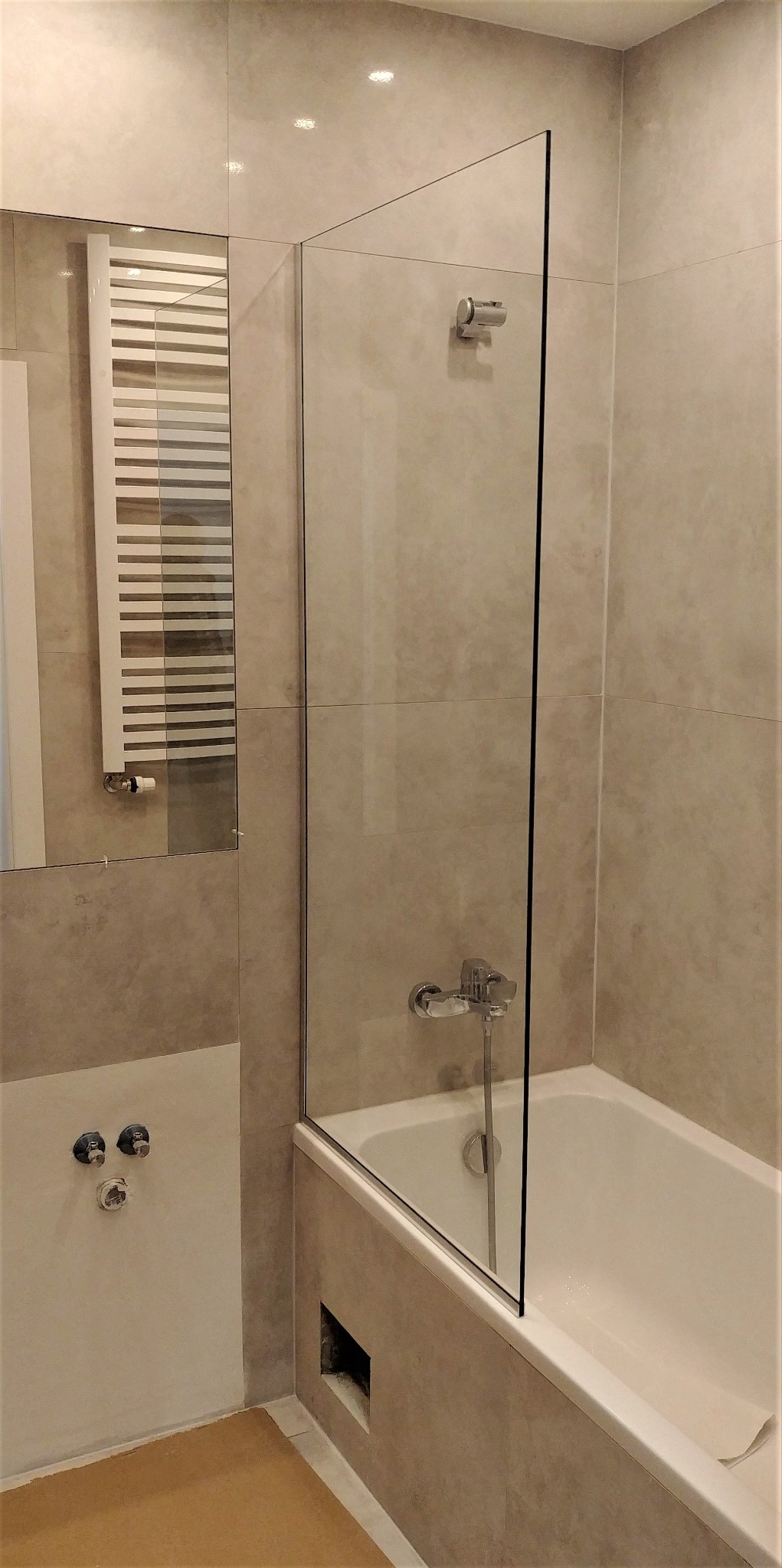 Szklana ścianka prysznicowa zamontowana na wannie w łazience wykończonej beżowymi płytkami, widoczny grzejnik drabinkowy i bateria wannowa.