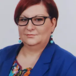 Portret kobiety w okularach, z rudymi włosami, ubranej w niebieską marynarkę i kolorową bluzkę, z turkusowymi kolczykami.