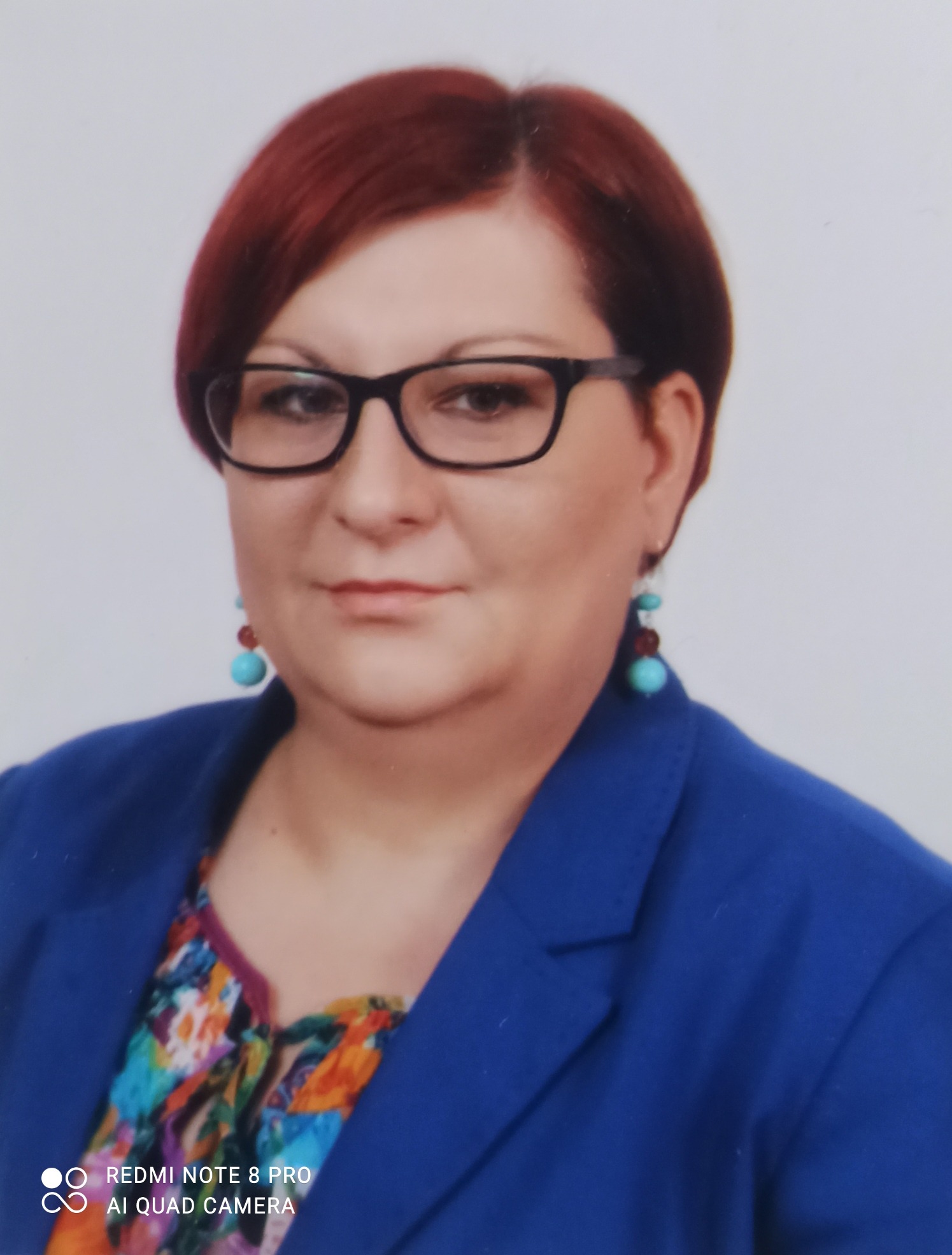 Portret kobiety w okularach, z rudymi włosami, ubranej w niebieską marynarkę i kolorową bluzkę, z turkusowymi kolczykami.