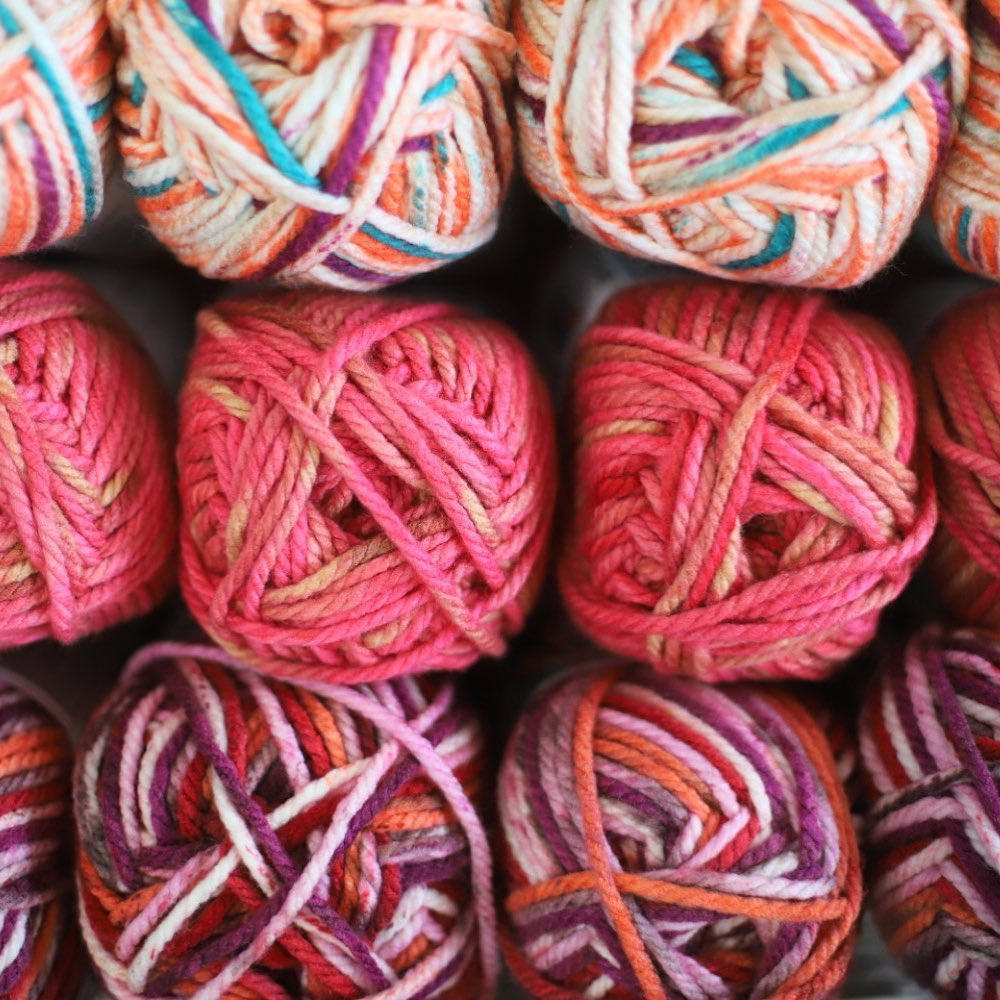 Kłębek kolorowej włóczki w odcieniach różu, pomarańczy, fioletu i bieli, ułożone w rzędach, widok z góry.