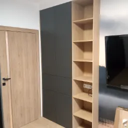 Nowoczesna szafa wnękowa z szarymi drzwiami i otwartą półką z jasnego drewna, obok drewnianych drzwi wewnętrznych.