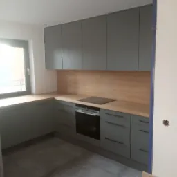 Szara zabudowa kuchenna z drewnianym blatem i płytą indukcyjną we Wrocławiu, widok na fragment wnętrza z szarymi płytkami podłogowymi.