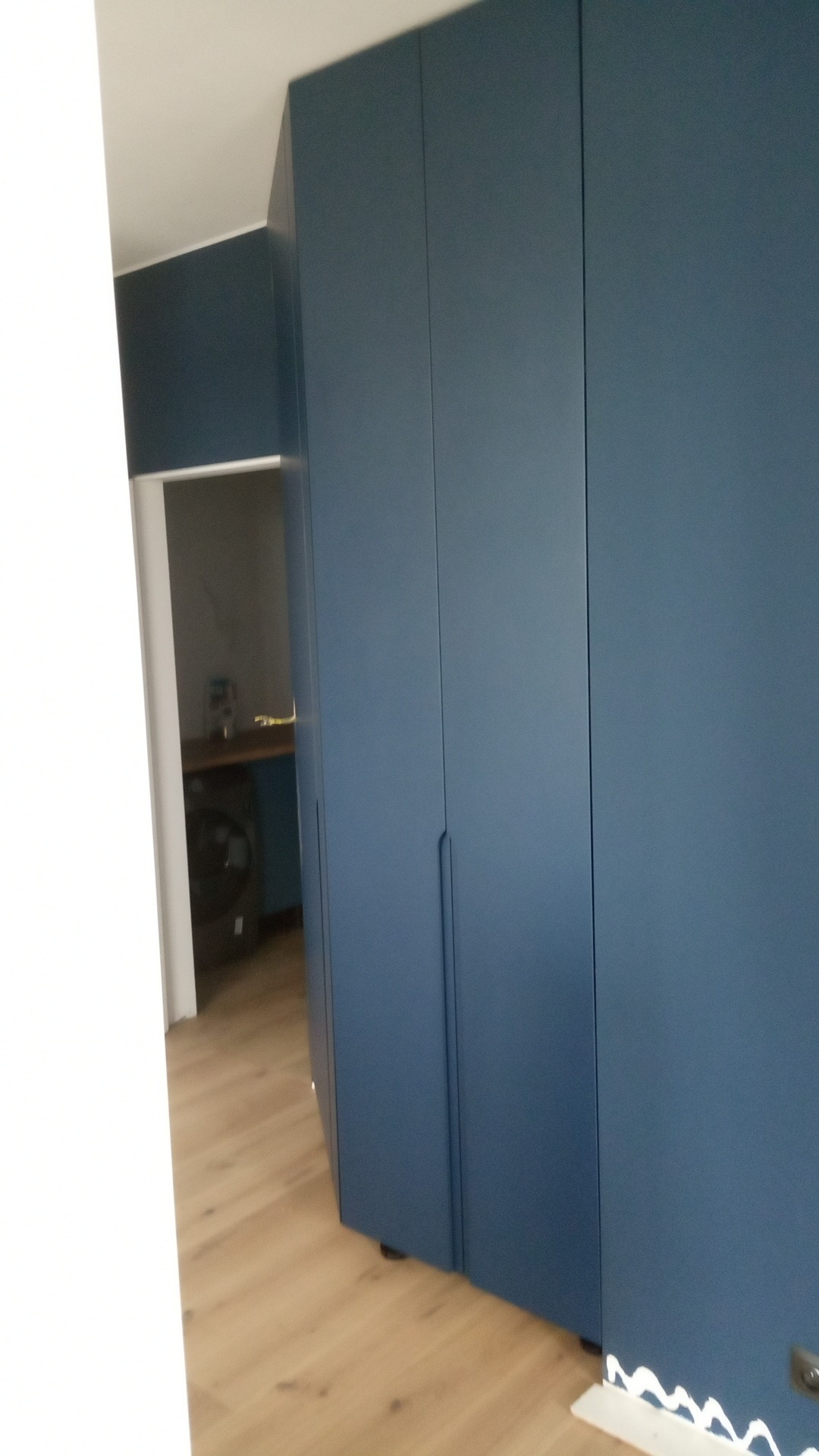 Niebieska szafa wnękowa z długimi, minimalistycznymi uchwytami, widoczna z boku, z fragmentem wnętrza pokoju z pralką w tle.