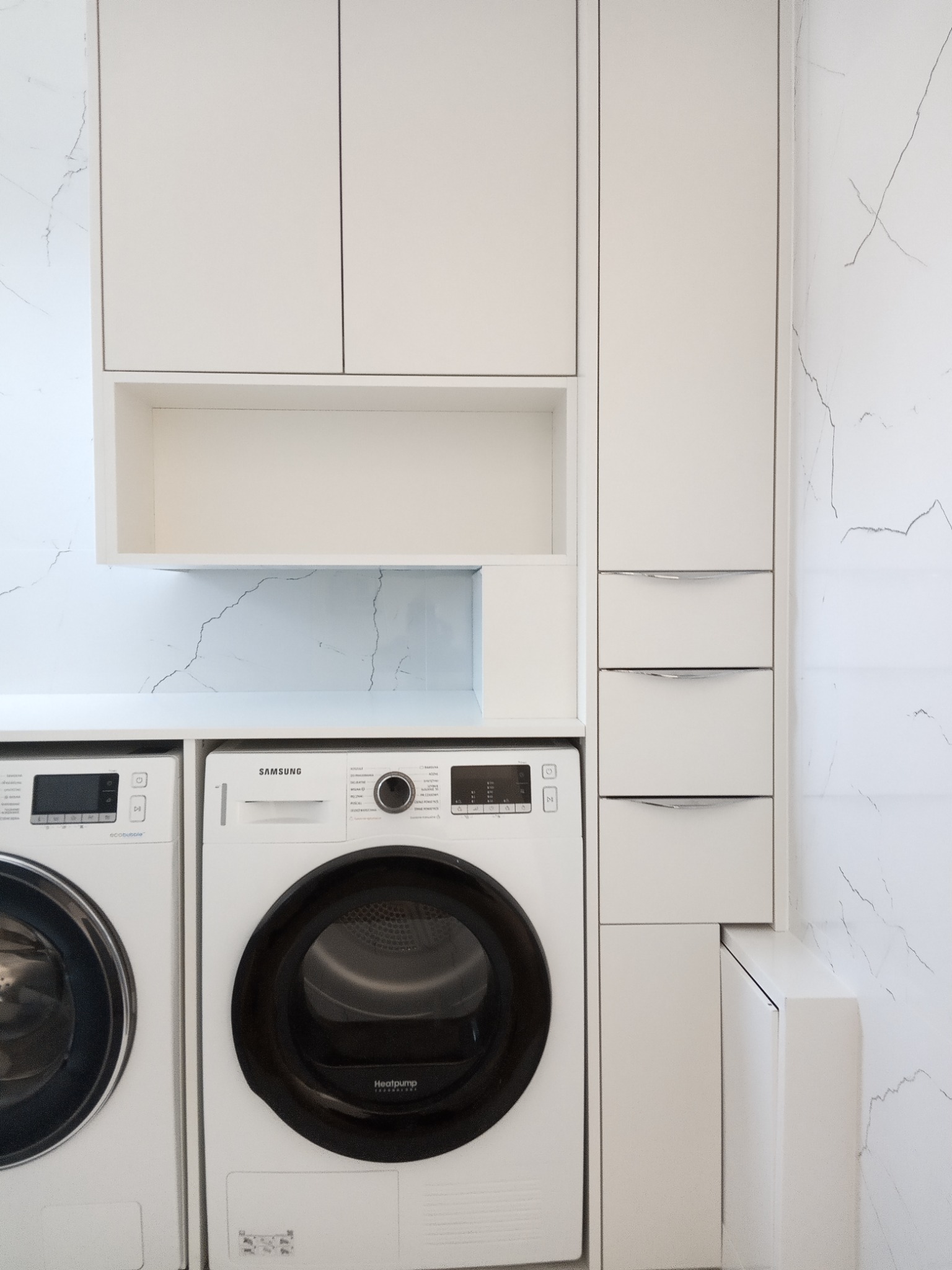 Biała zabudowa meblowa w pralni z pralką i suszarką Samsung, marmurowe płytki na ścianie.
