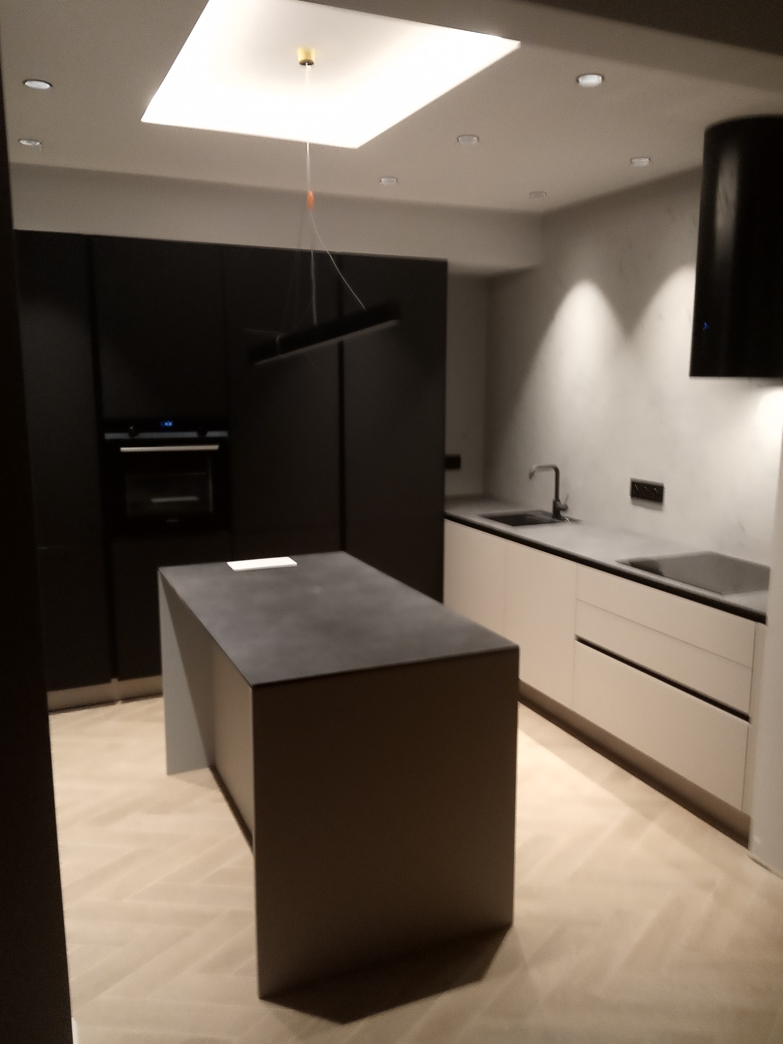 Nowoczesna kuchnia z wyspą w odcieniach szarości i bieli, oświetlona wpuszczanymi lampami i wiszącą lampą nad wyspą, z parkietem ułożonym w jodełkę.