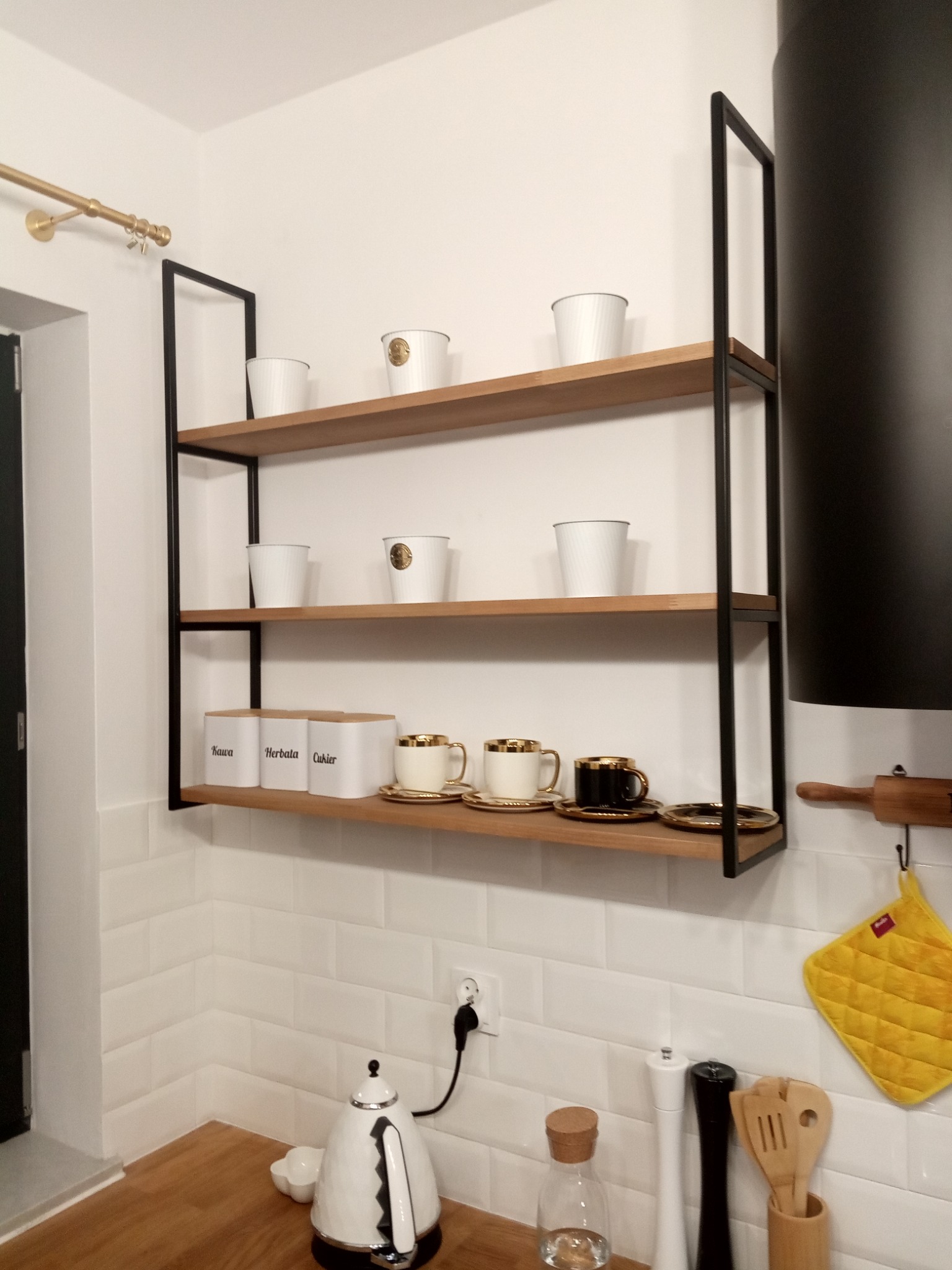 Półka wisząca z drewnianymi blatami i czarną metalową ramą, na której stoją białe pojemniki z napisami Kawa, Herbata, Cukier oraz filiżanki w kolorze białym i czarnym ze złotymi zdobieniami, w tle...