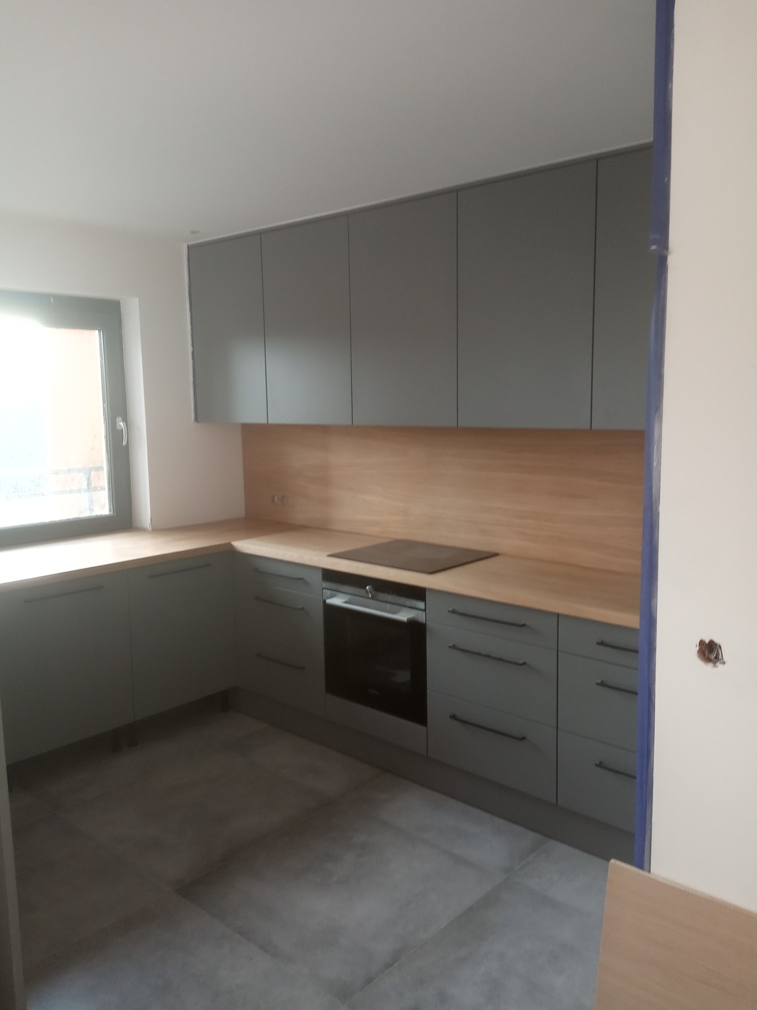Szara zabudowa kuchenna z drewnianym blatem i płytą indukcyjną we Wrocławiu, widok na fragment wnętrza z szarymi płytkami podłogowymi.