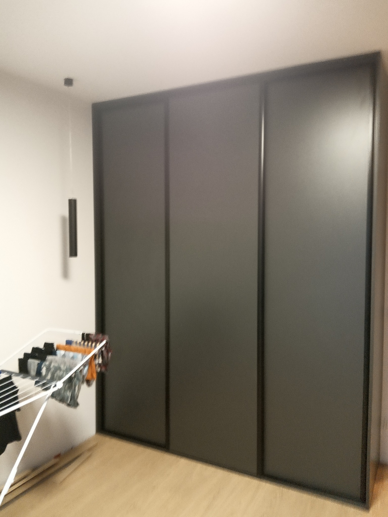 Trzydrzwiowa szafa przesuwna w kolorze grafitowym z czarnymi obramowaniami, widoczna suszarka na pranie w rogu pokoju.