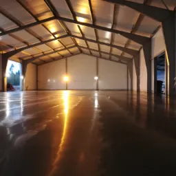 Błyszcząca, ciemna podłoga w hali magazynowej z metalową konstrukcją dachu i oświetleniem, widoczne odbicia świateł w posadzce.