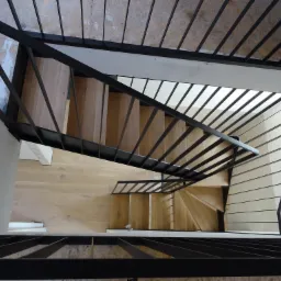 Widok z góry na schody z drewnianymi stopniami i czarną, minimalistyczną balustradą z metalowych prętów, z perspektywy pionowej.