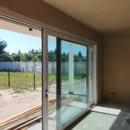 Wnętrze w stanie deweloperskim z widokiem na zewnątrz przez przesuwne okno tarasowe; ściany przygotowane do malowania, podłoga z wylewką, na zewnątrz widoczne drzewa i ogrodzenie.