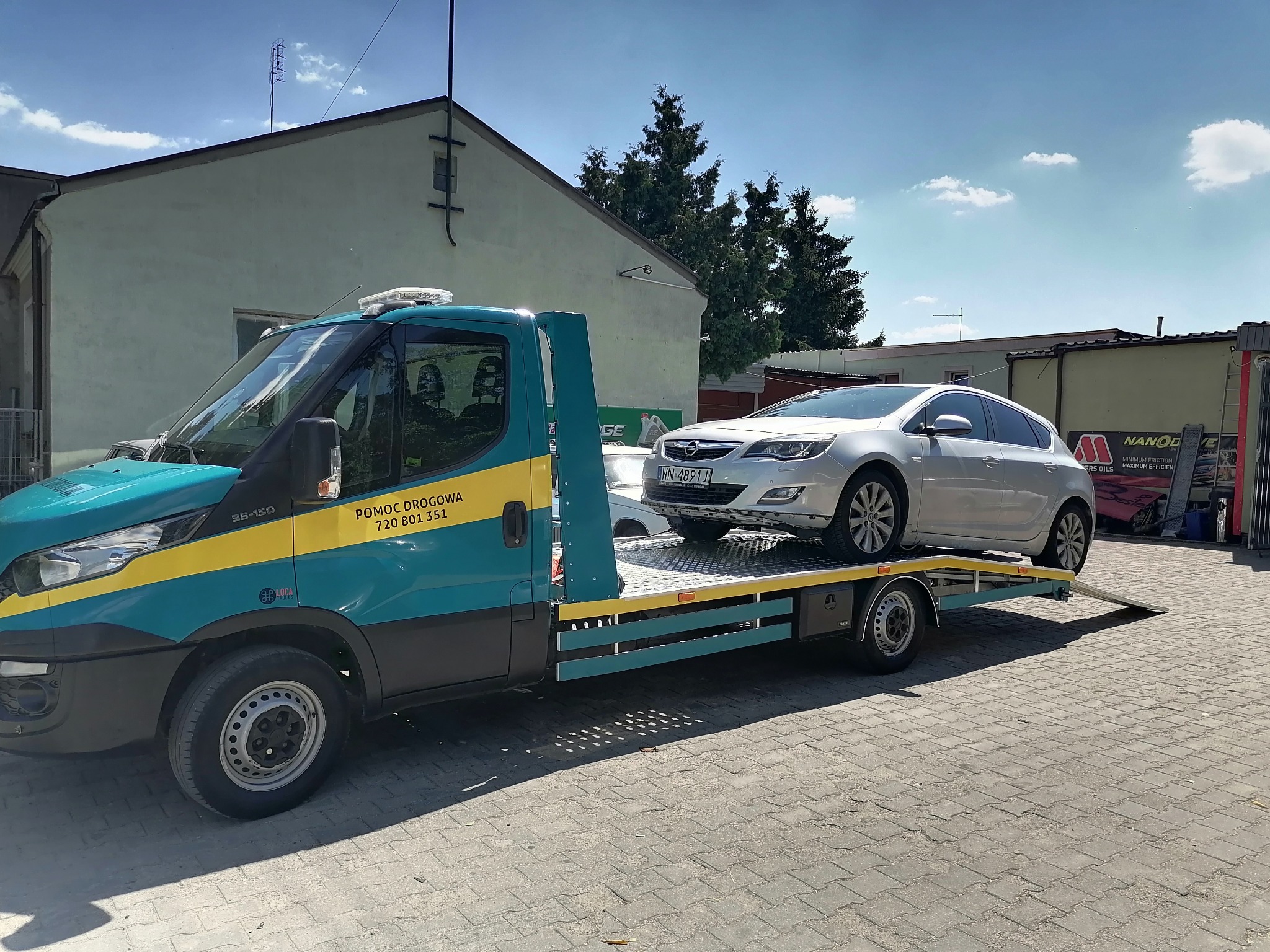 Turkusowa ciężarówka pomocy drogowej z żółtym pasem, przewożąca srebrnego Opla Astrę na metalowej platformie, zaparkowana na brukowanym placu przed budynkiem.