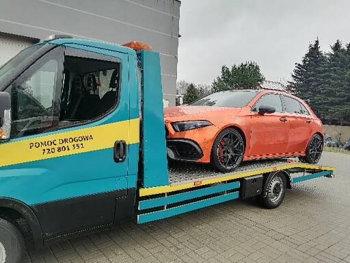 Pomarańczowy samochód osobowy marki Mercedes-Benz klasy A transportowany na niebiesko-żółtej platformie pojazdu pomocy drogowej z napisem 'Pomoc Drogowa 720 801 351'.