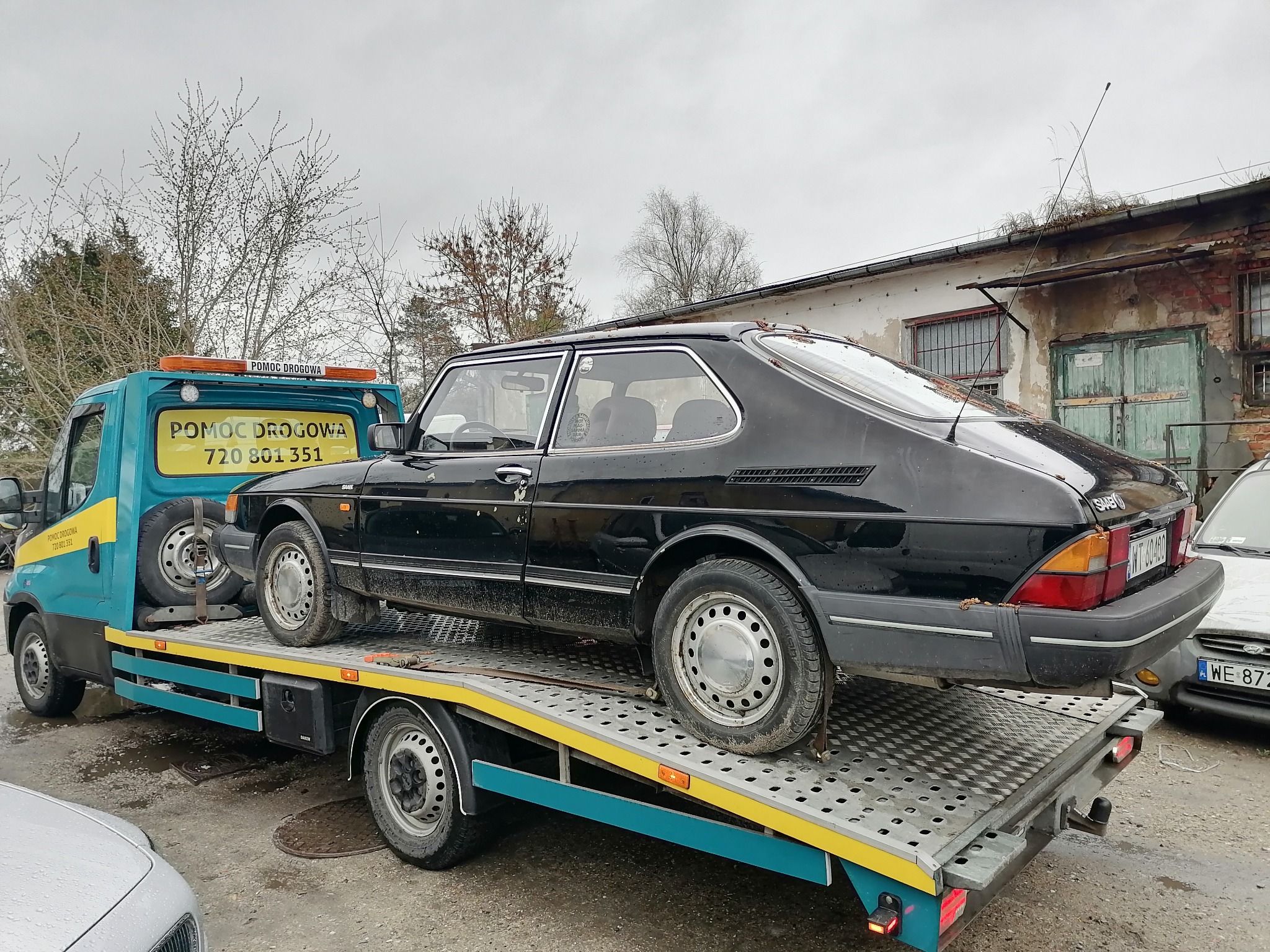 Czarny Saab 900 na platformie pomocy drogowej z napisem 'POMOC DROGOWA 720 801 351' w tle budynek z zielonymi drzwiami i anteną na dachu.