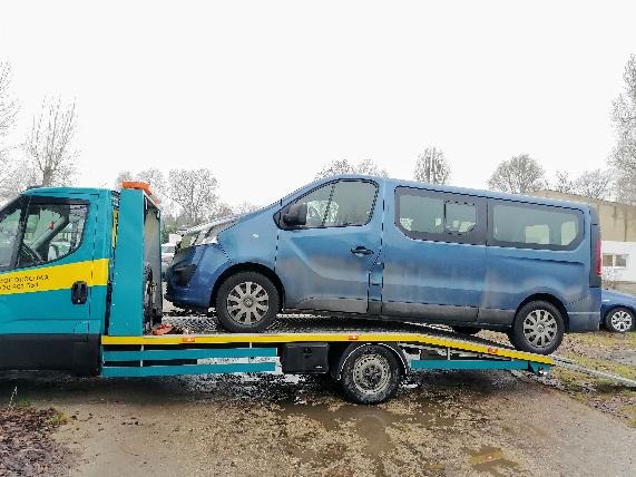 Niebieski van na platformie pojazdu holowniczego, widoczne uszkodzenia karoserii z przodu, mokra nawierzchnia placu.