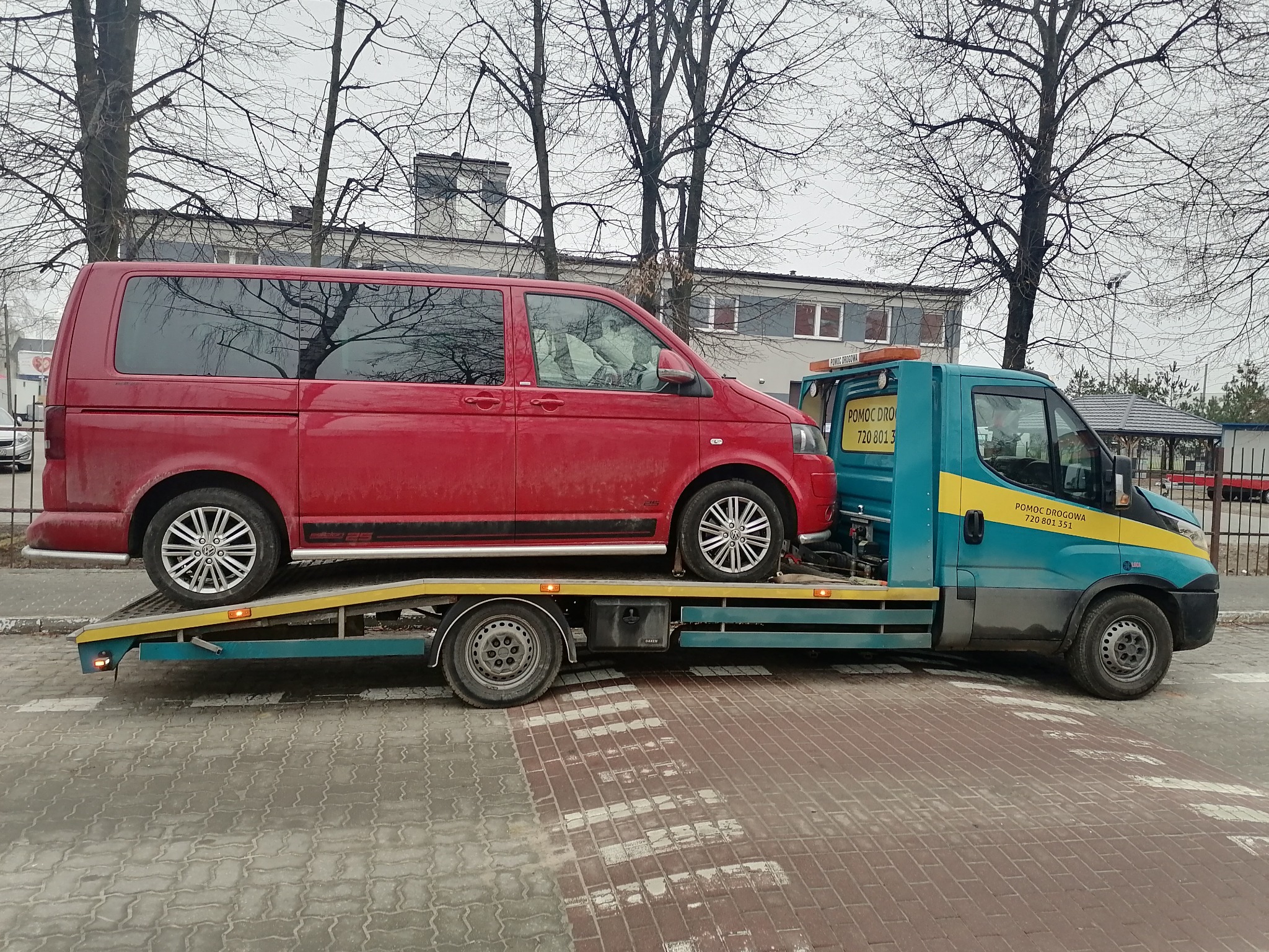 Czerwony van Volkswagen T5 Edition 25 na platformie transportowej niebiesko-żółtej pomocy drogowej na tle miejskiego krajobrazu z drzewami bez liści.