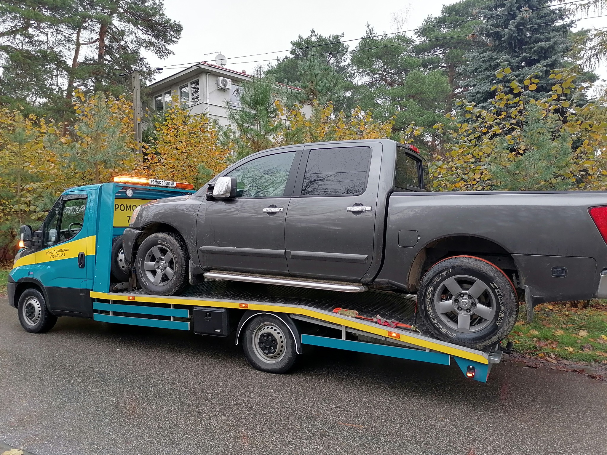 Laweta w kolorach turkusowym i żółtym transportuje szarego pickupa Nissan Titan na tle jesiennego krajobrazu z domem w oddali.