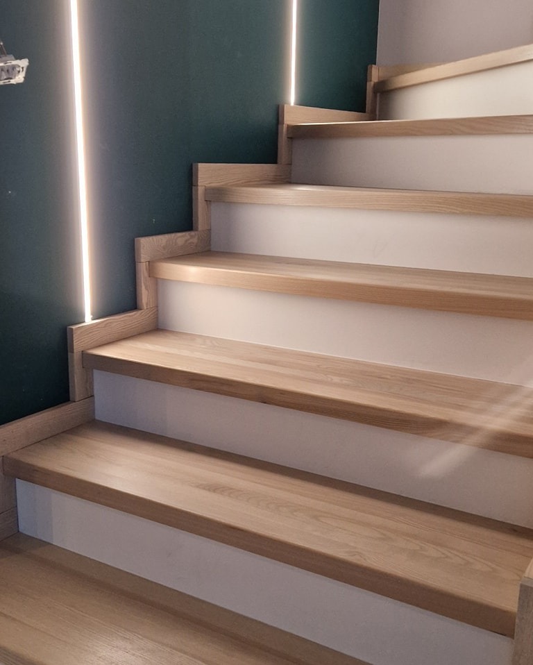 Jasne, dębowe stopnie schodów z podświetleniem LED na ciemnozielonej ścianie, widok z dołu.