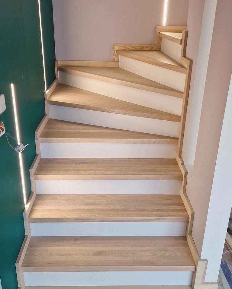 Jasne, drewniane schody z białymi podstopnicami, oświetlenie LED wzdłuż ściany, widoczne okablowanie elektryczne.