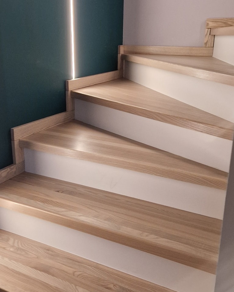 Jasne drewniane schody z białymi podstopnicami, oświetlenie LED wzdłuż ściany, minimalistyczny design.