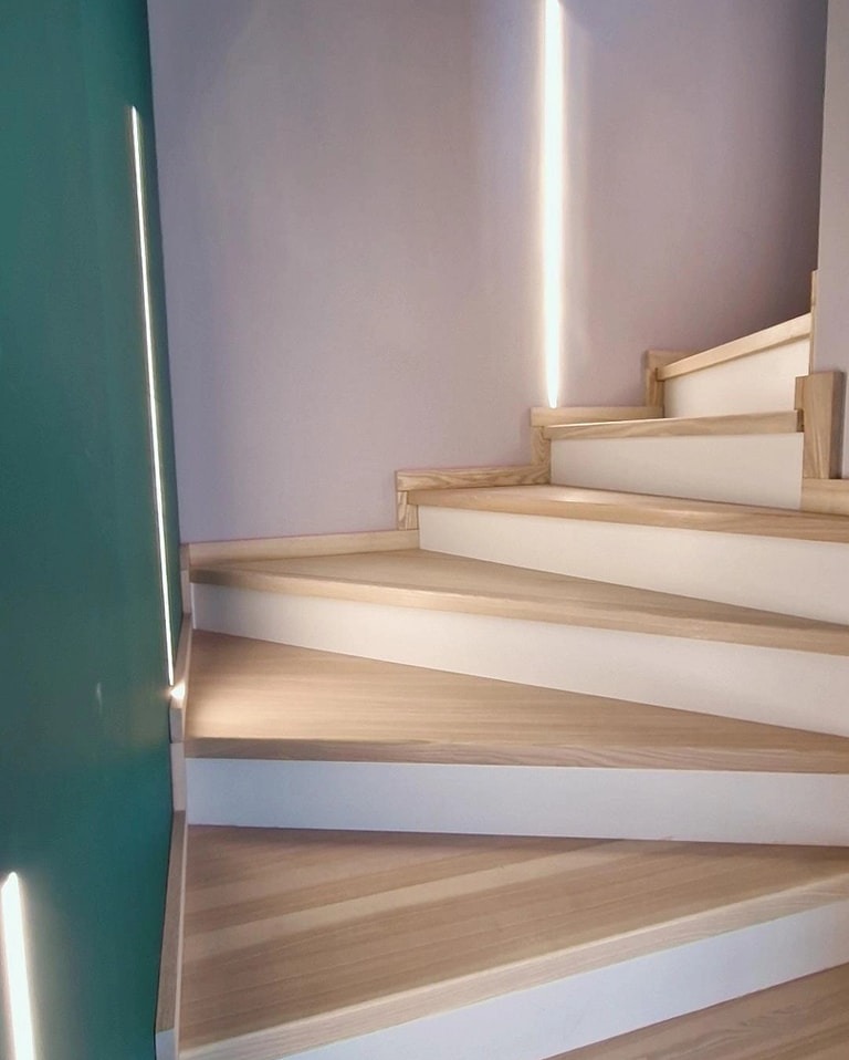 Nowoczesne schody z jasnego drewna z białymi podstopnicami, oświetlenie LED w ścianie, widok z dołu.