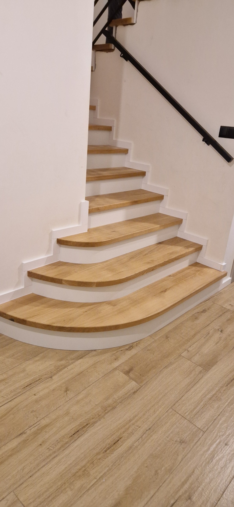 Bieg schodów z drewnianymi stopniami o zaokrąglonych krawędziach, osadzonych na białej konstrukcji, z czarną metalową poręczą i podłogą imitującą drewno.