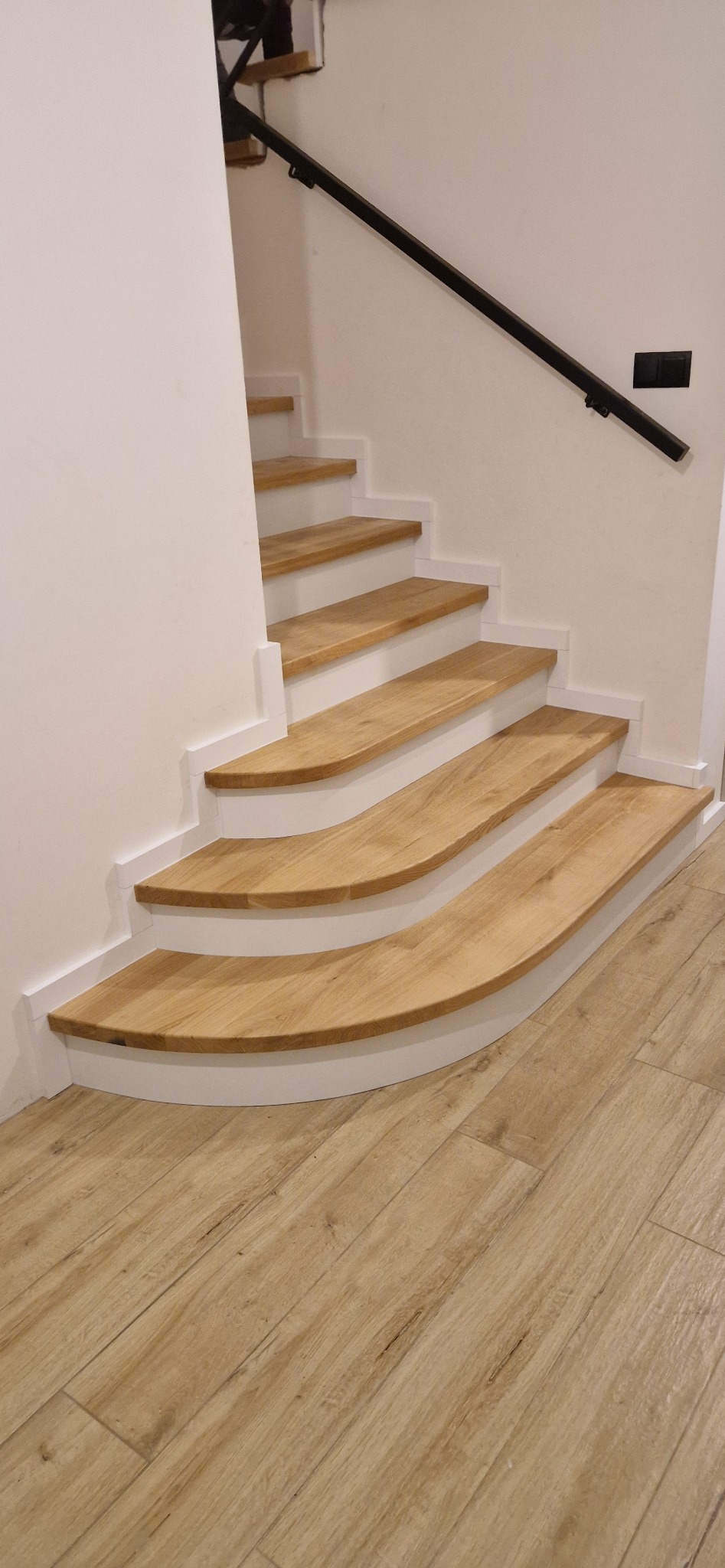 Nowoczesne schody z drewnianymi stopniami o zaokrąglonych krawędziach, wykończone białymi podstopnicami i czarną, minimalistyczną balustradą, harmonizujące z podłogą imitującą drewno.