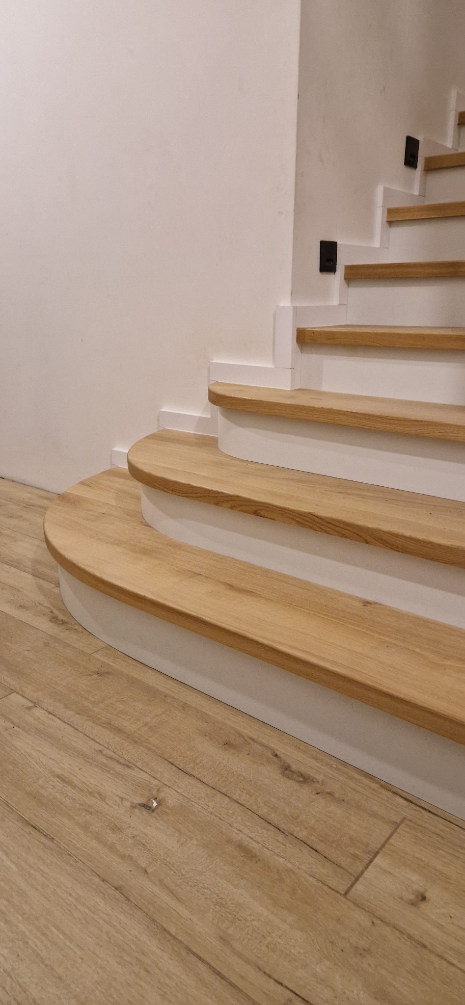 Fragment schodów z drewnianymi stopniami o zaokrąglonym kształcie, pomalowanymi na jasny odcień dębu, z białymi podstopnicami i minimalistycznymi, czarnymi uchwytami, widoczne na tle podłogi...