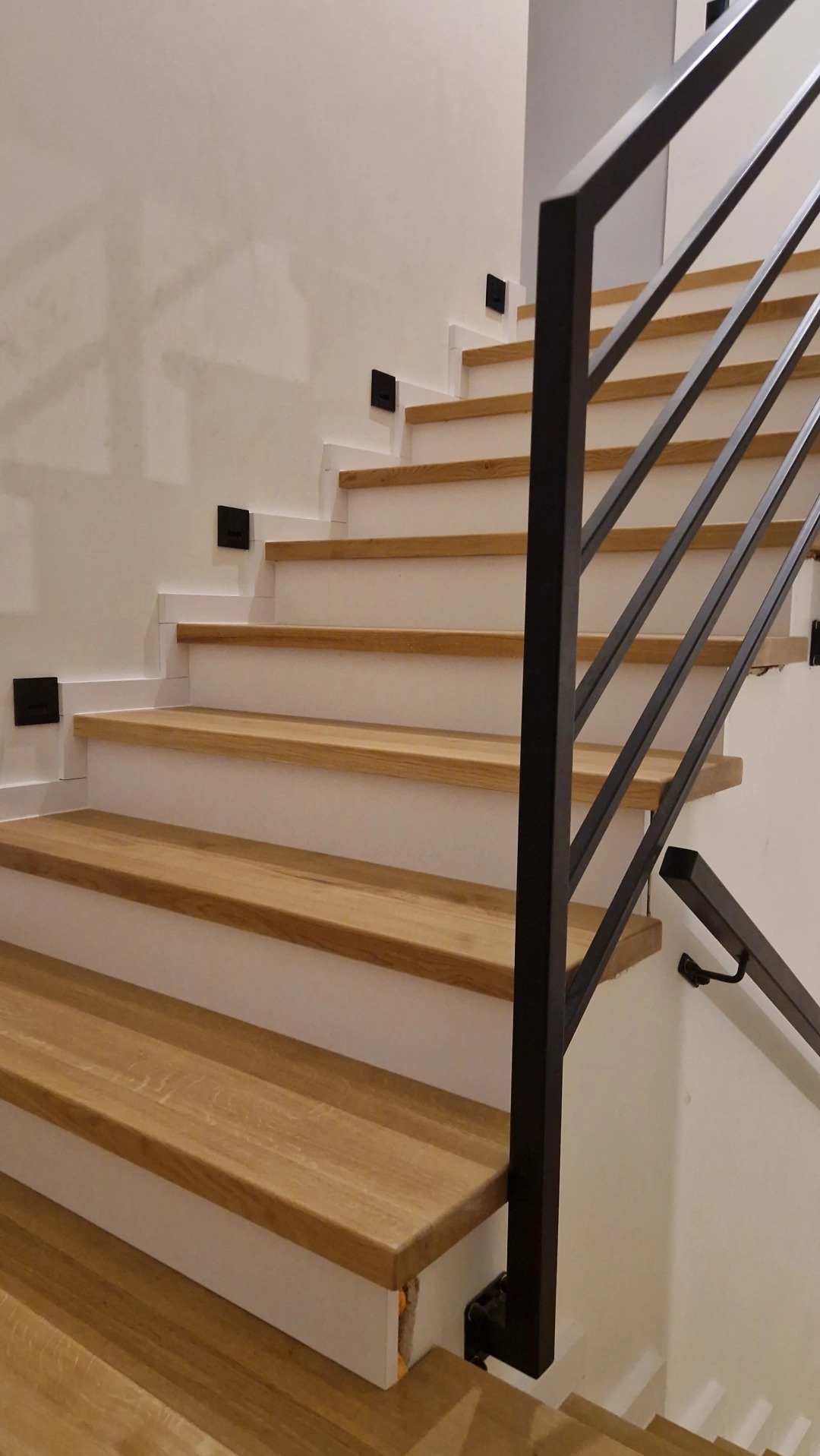 Nowoczesne schody z drewnianymi stopniami i czarną, metalową balustradą, oświetlone punktowymi lampkami przy ścianie.